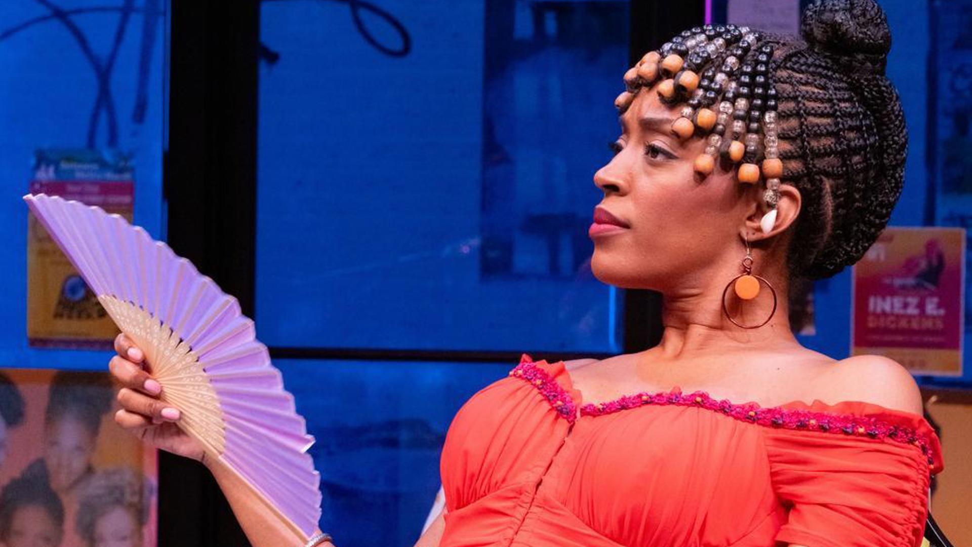 A performer sits in a salon chair with a serious expression, fanning themselves with a hand fan.