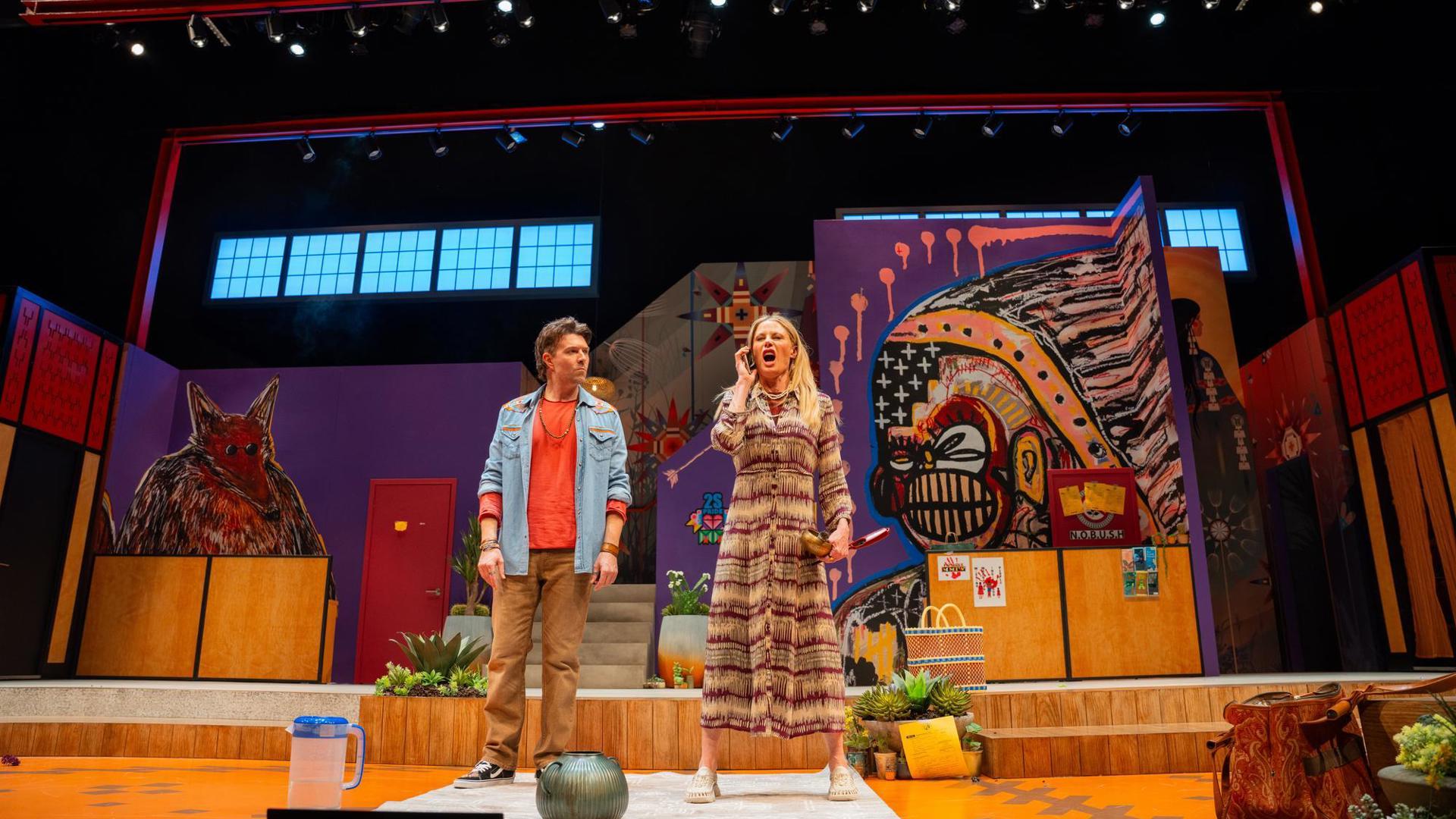 Two people stand on a colorful stage adorned with vibrant Indigenous-inspired artwork, one person is having a heated coversation on a phone the other stands there looking puzzled.