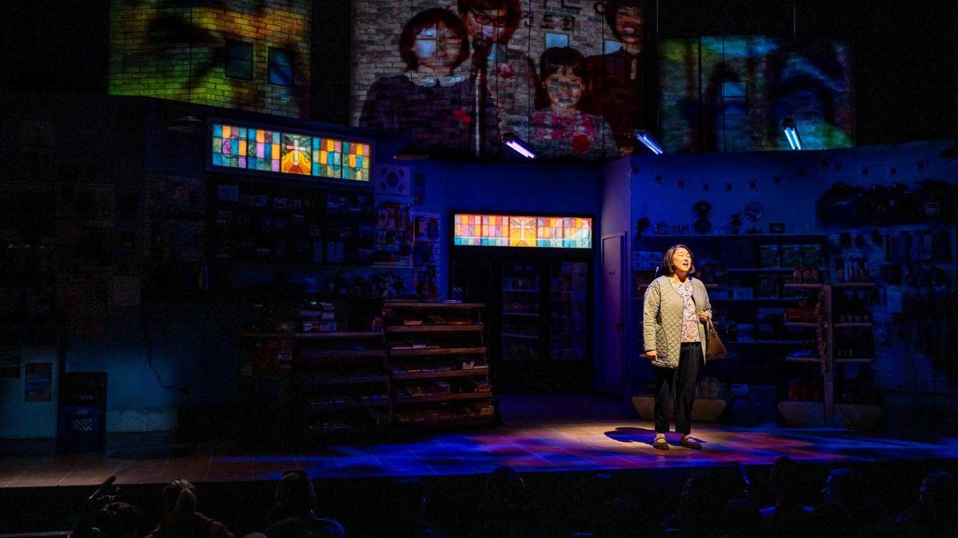 A woman stands downstage in a dark convenience store, projects of family portraits are seen on the wall. 