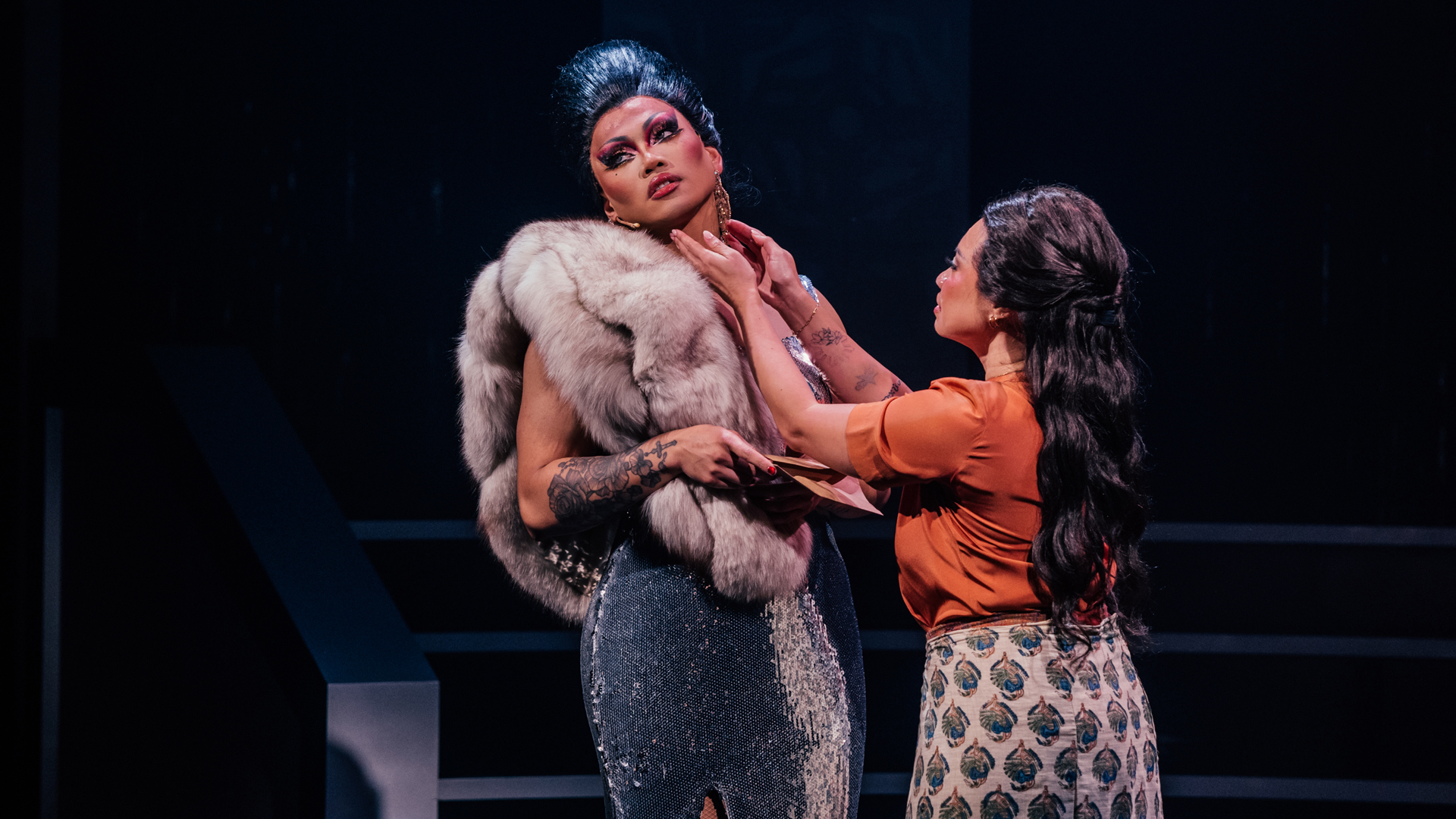 A performer reaches for a glamorously-dressed performer's face, who leans away from them.