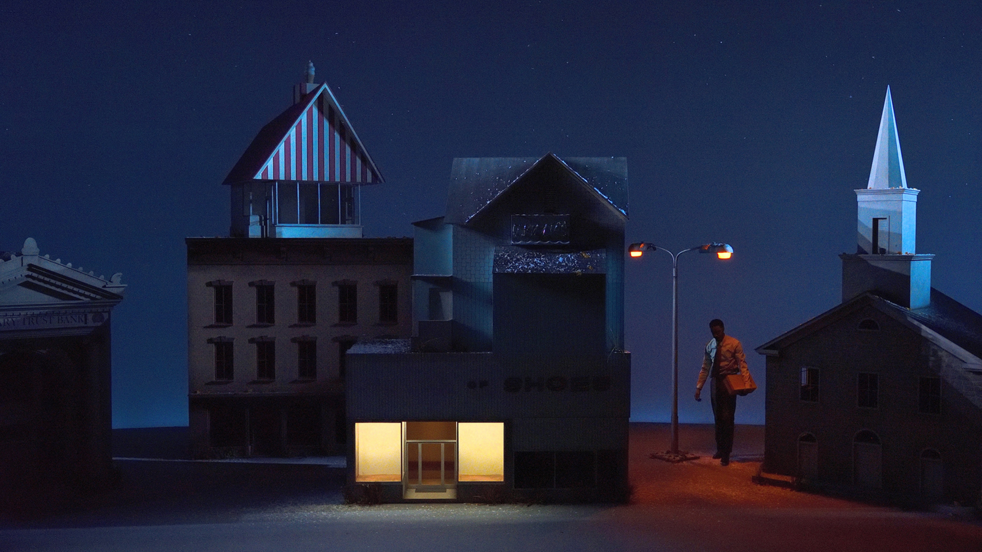 A man holding a shoebox stands under a lit streetlamp in a town, surrounded by large buildings.
