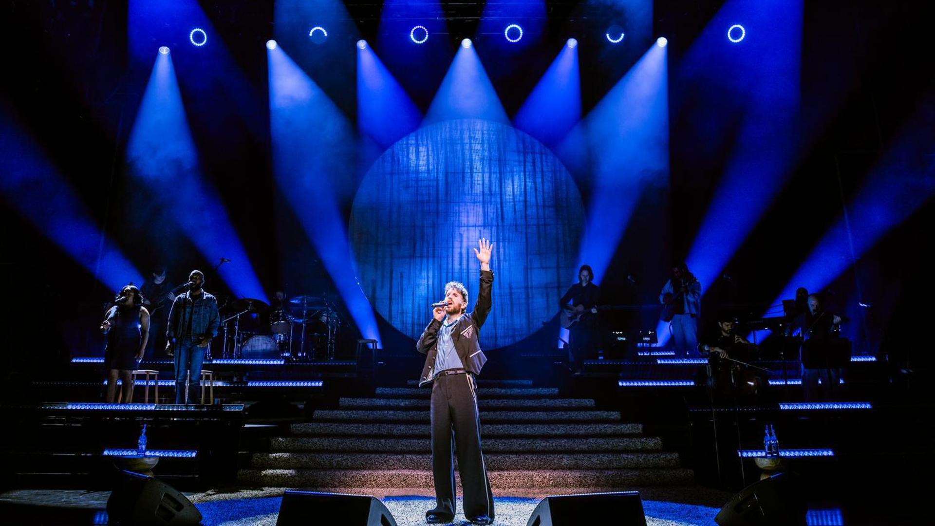 Ben Platt singing with a raised hand, blue stage lights fill the stage. 