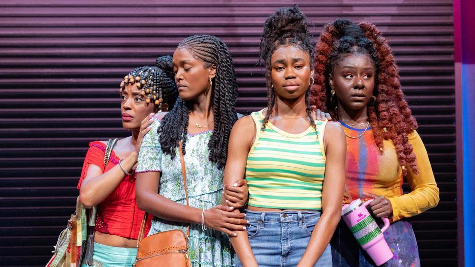 Four performers stand center stage embracing each other, with forlorn expressions.