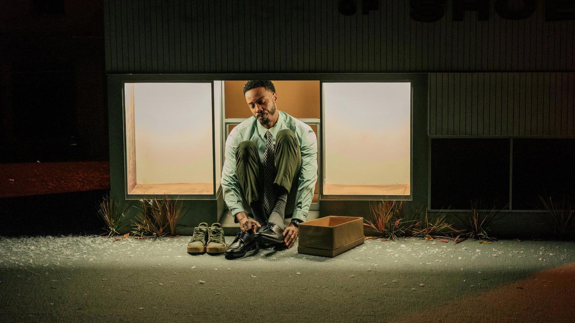 A man sits in front of a building. between two lit windows, trying on a pair of shoes.