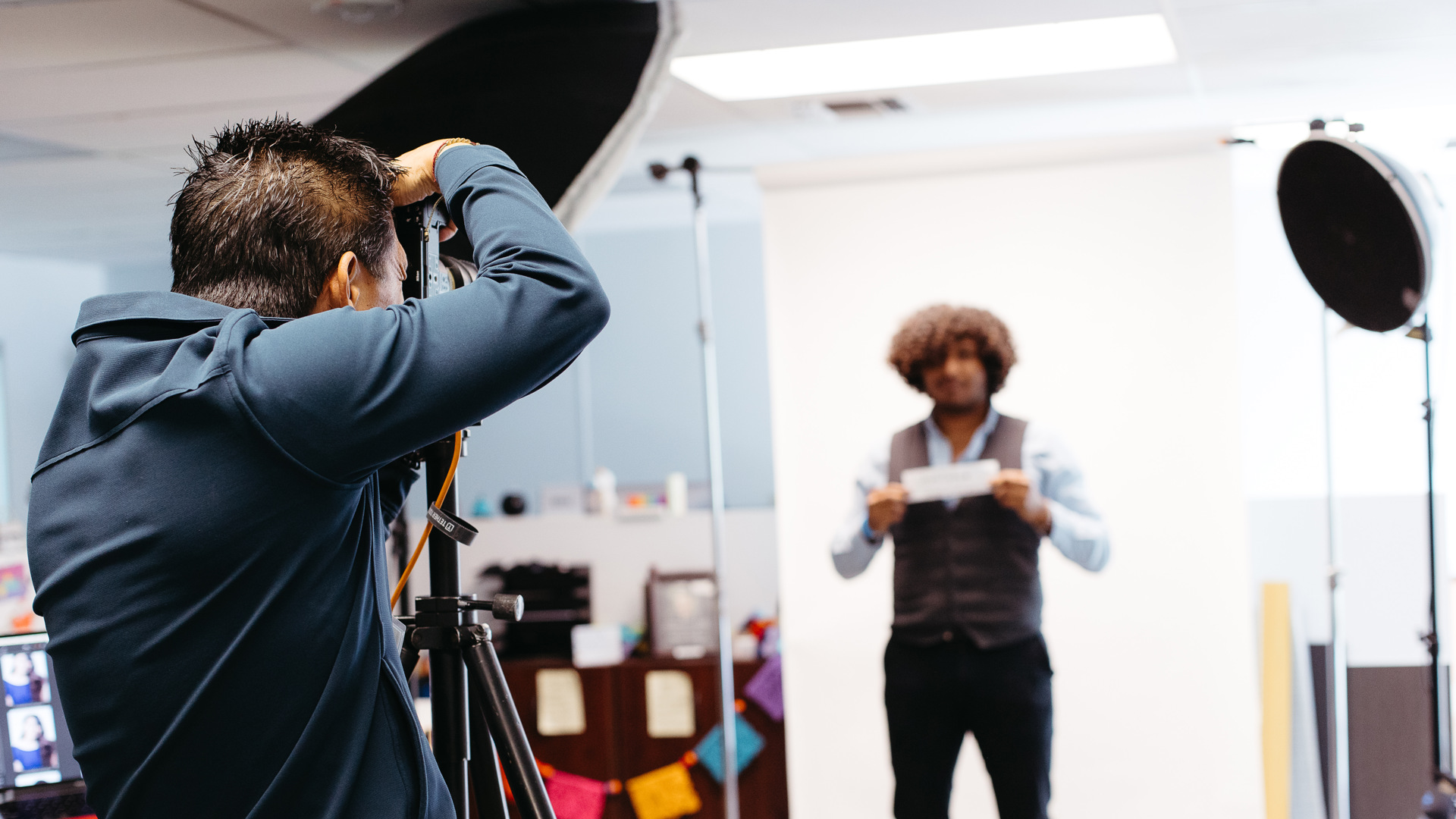 A photographer takes a headshot style photo of a person standing infront of a white backdrop. 