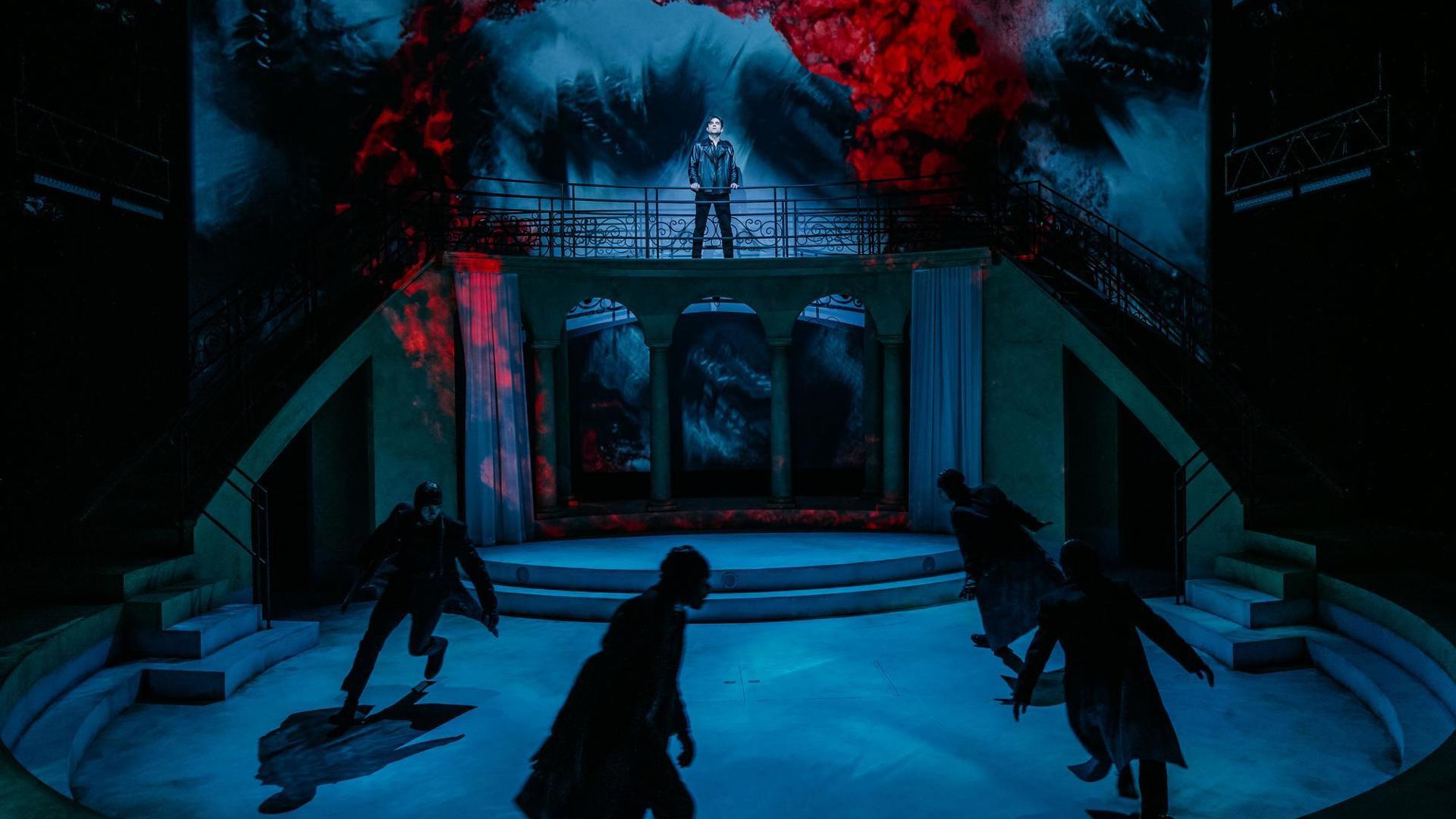 An actor stands on a platform, looking out to the audience. Beneath them, four ensemble members run in a circle.