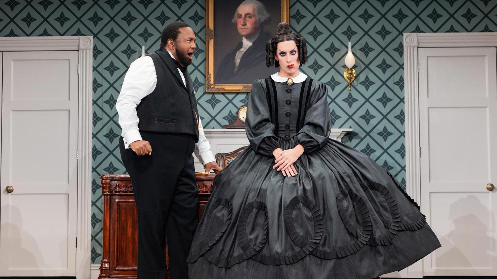 An actor in a black vest, a white shirt and black pants yells at an actress wearing a black dress, who looks out at the audience angrily.