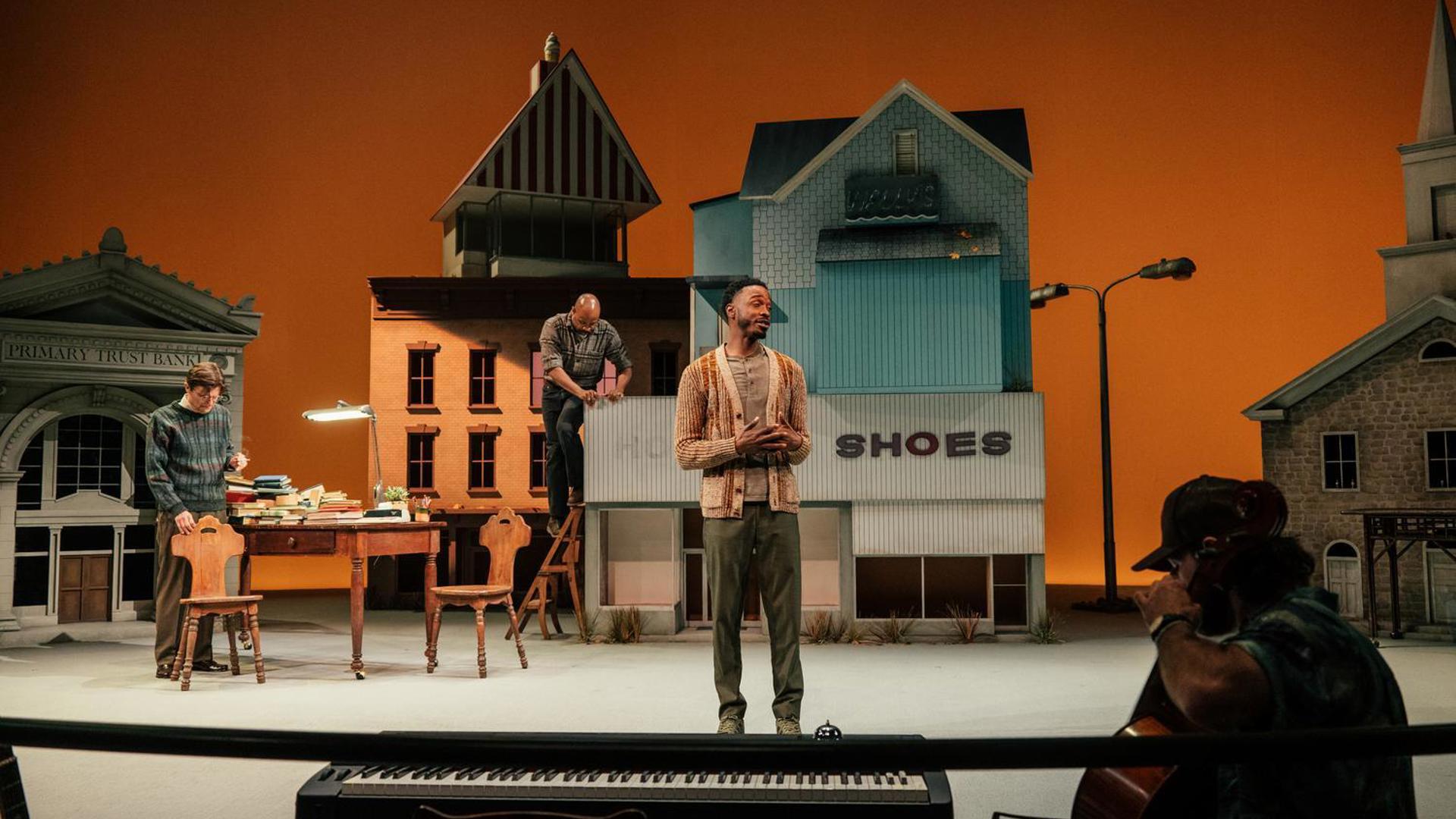 Three men stand on stage in front of large buildings, including a bank, a shoe store, and a church, while another man in the foreground sits in a chair beside a piano keyboard while playing the cello.
