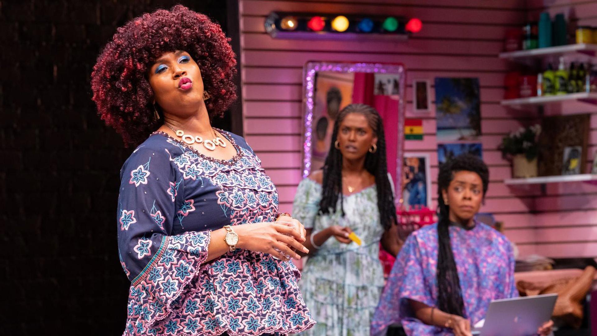 A performer stands in the foreground, speaking. Two performers watch them from the background.