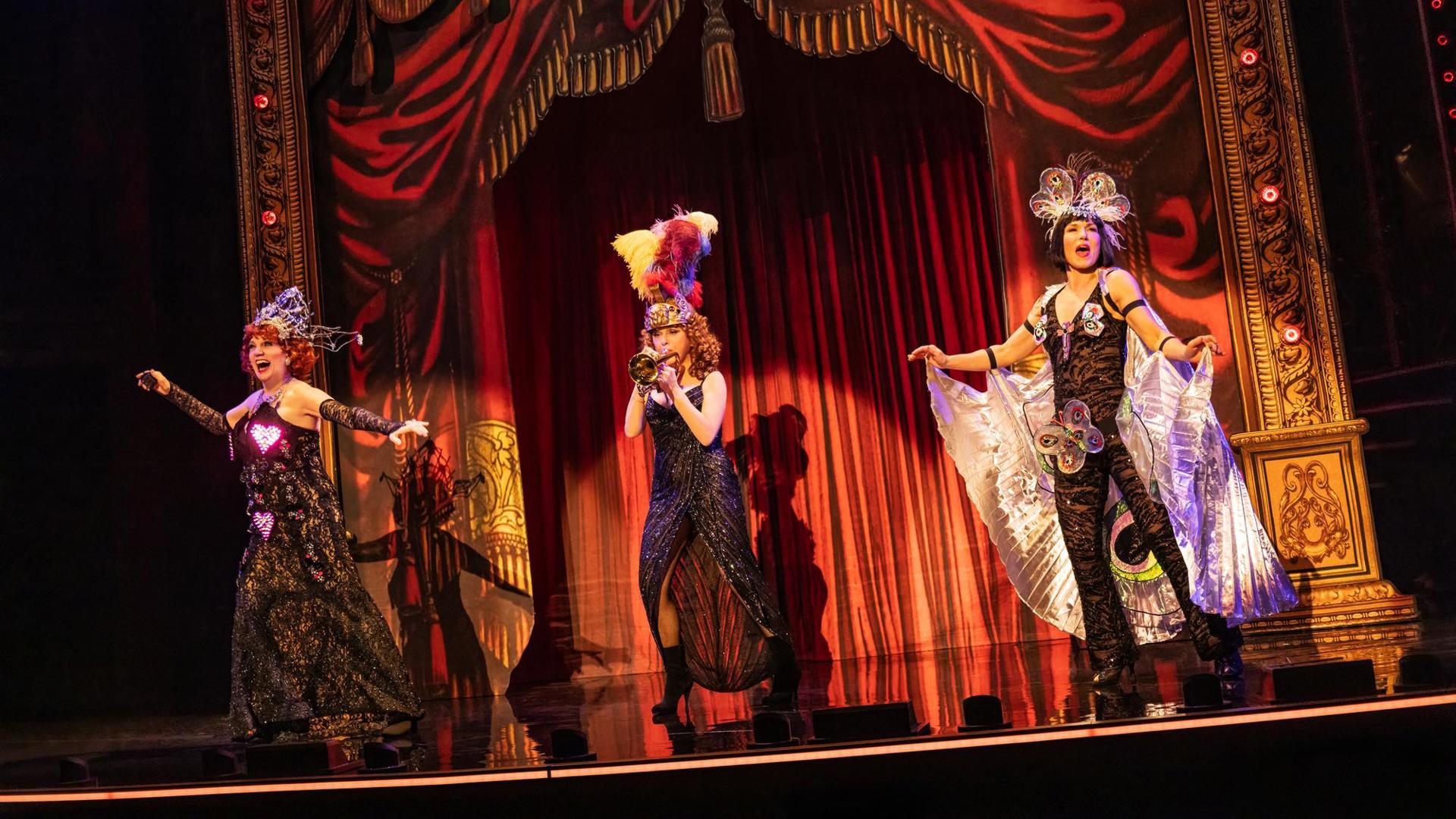 Three performers, wearing flashy costumes, pose onstage. One performer play a trumpet.