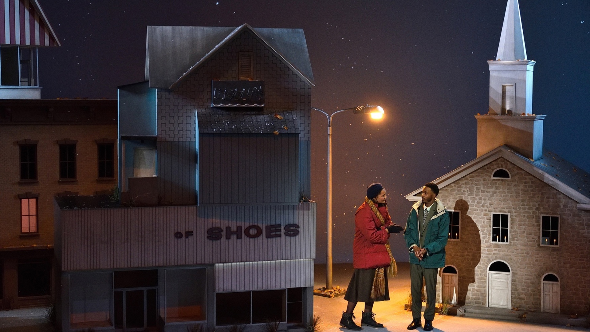 A woman and man dressed in winter coats stand underneath a streetlamp in front of a church in a town full of large buildings as snow falls from above.