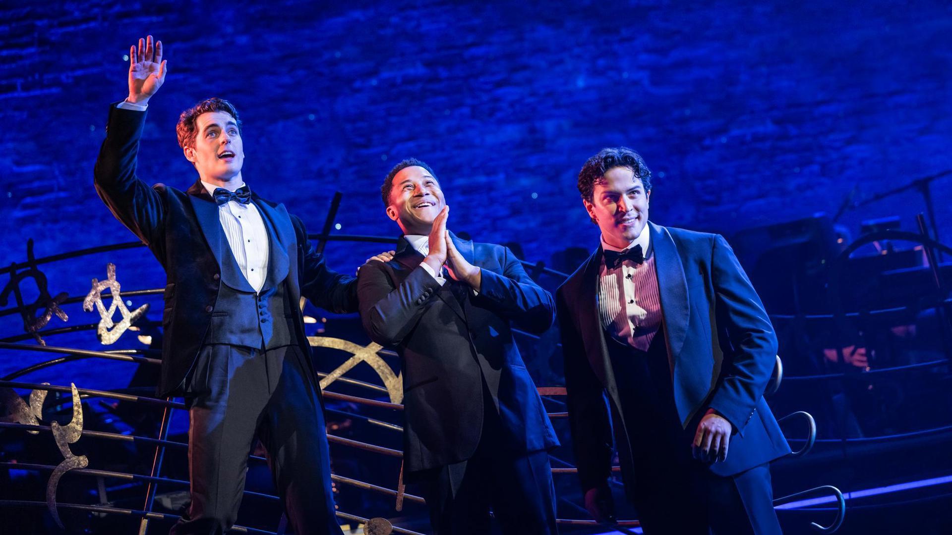 Three performers wear tuxedos and look out toward the audience. One raises their hand, another clasps their hands together.