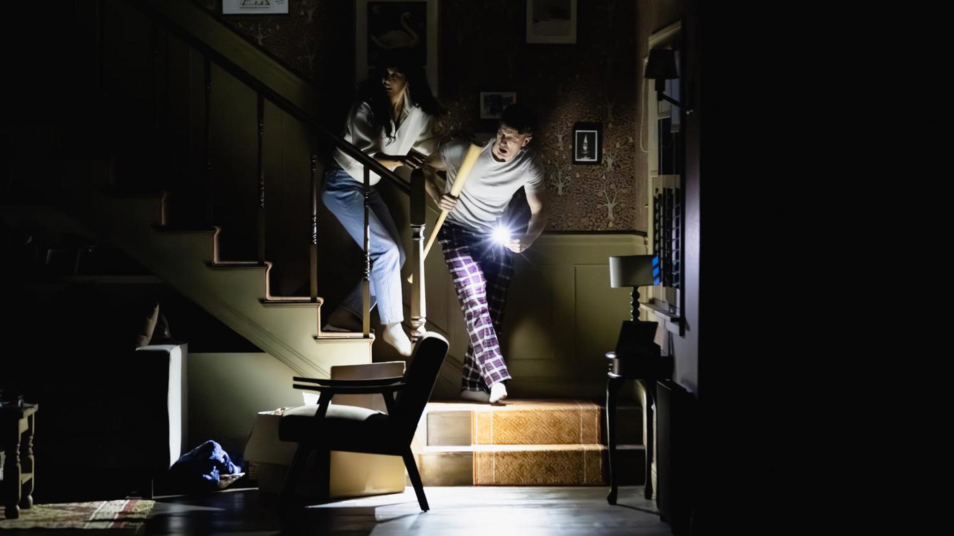 A couple coming down the stairs with a flashlight and a baseball bat.