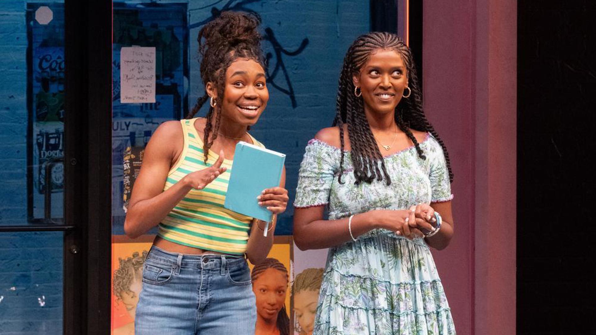 Two performers stand center stage. The performer on the left speaks while the performer on the right listens.