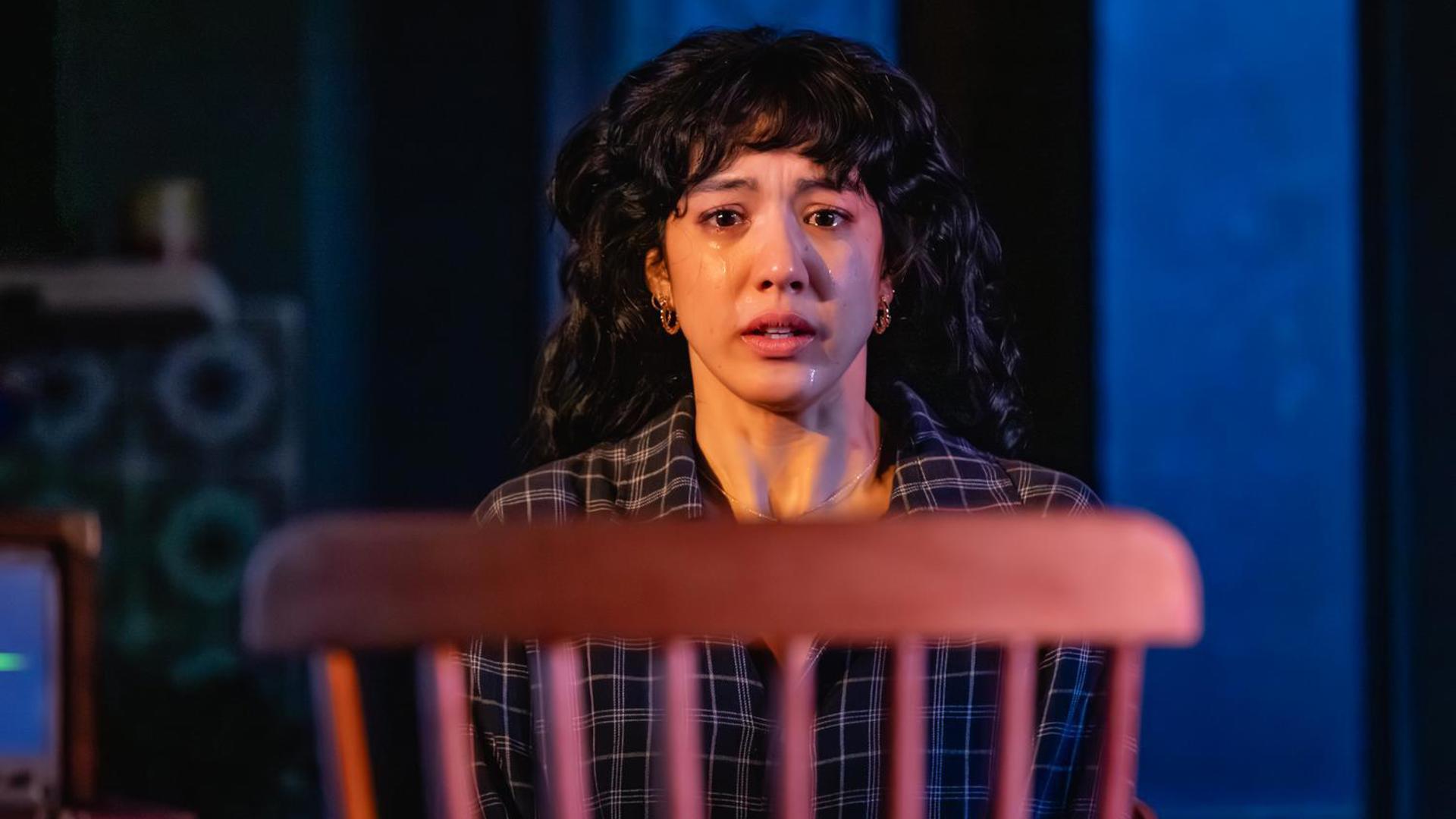 A woman crying while looking at a chair in the foreground.