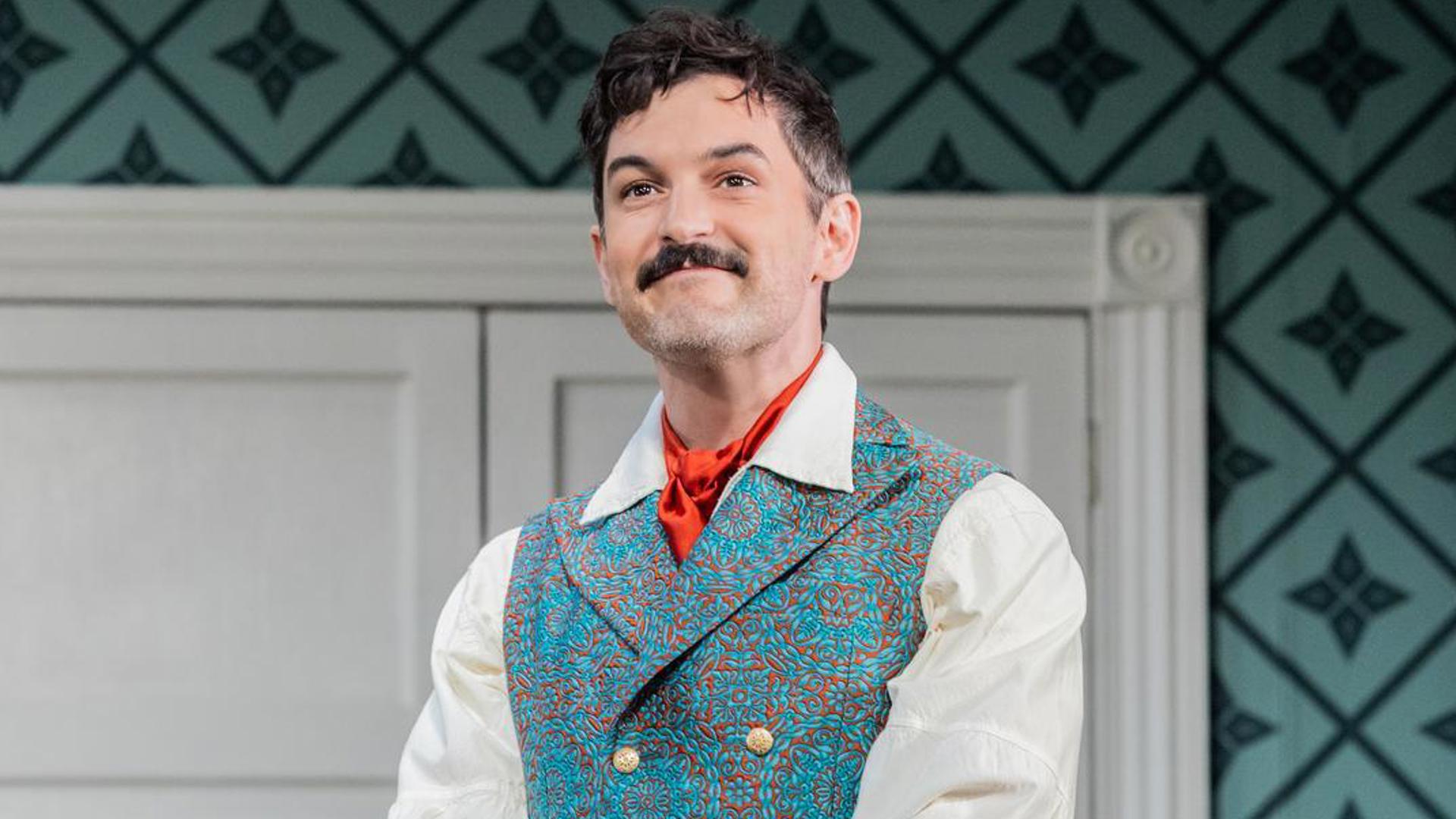 An actor stands in a blue vest, looking out at the audience smiling.