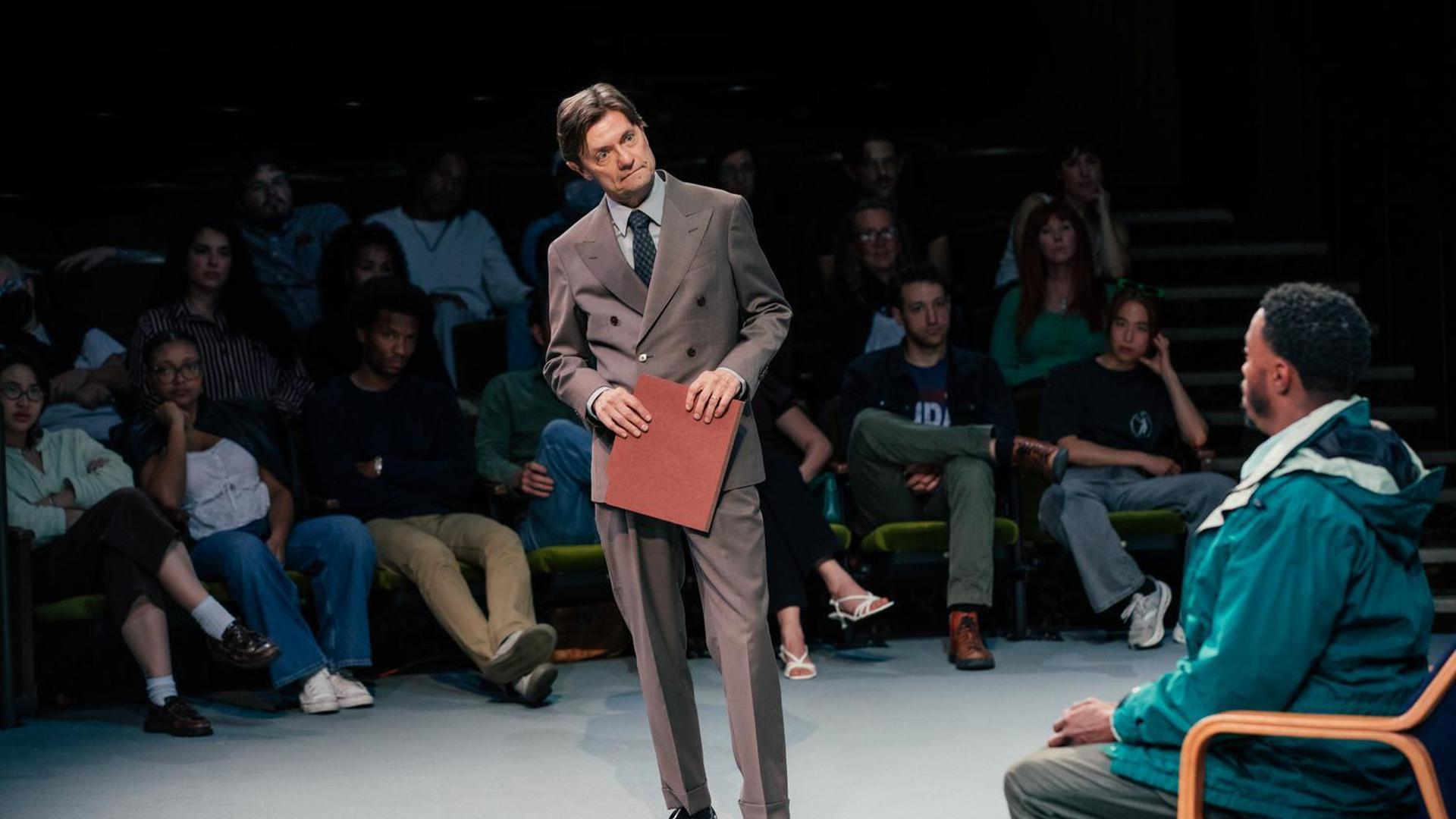 A man in a suit and tie holds a folder and is looking the direction of another man who sits in a chair looking back at him.
