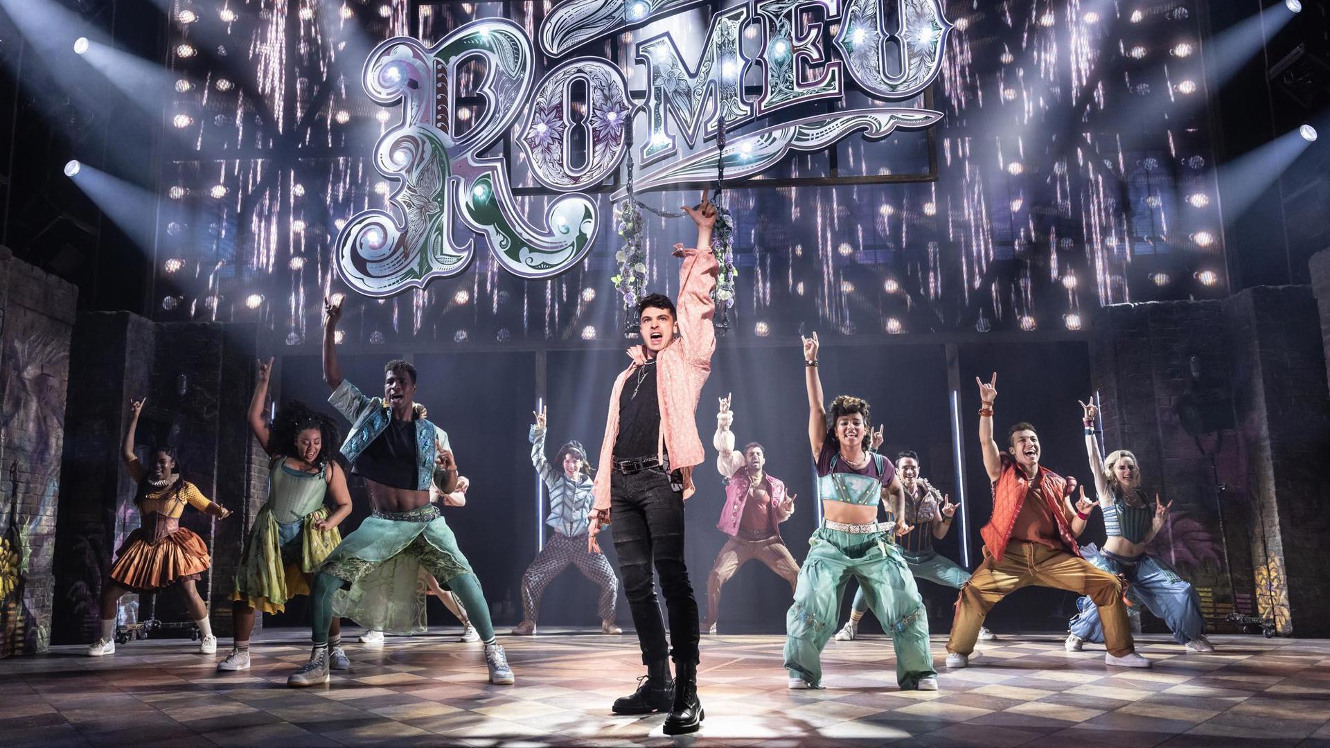 Michael Canu poses center stage beneath a large sing that reads "Romeo" while dancers dance behind him.